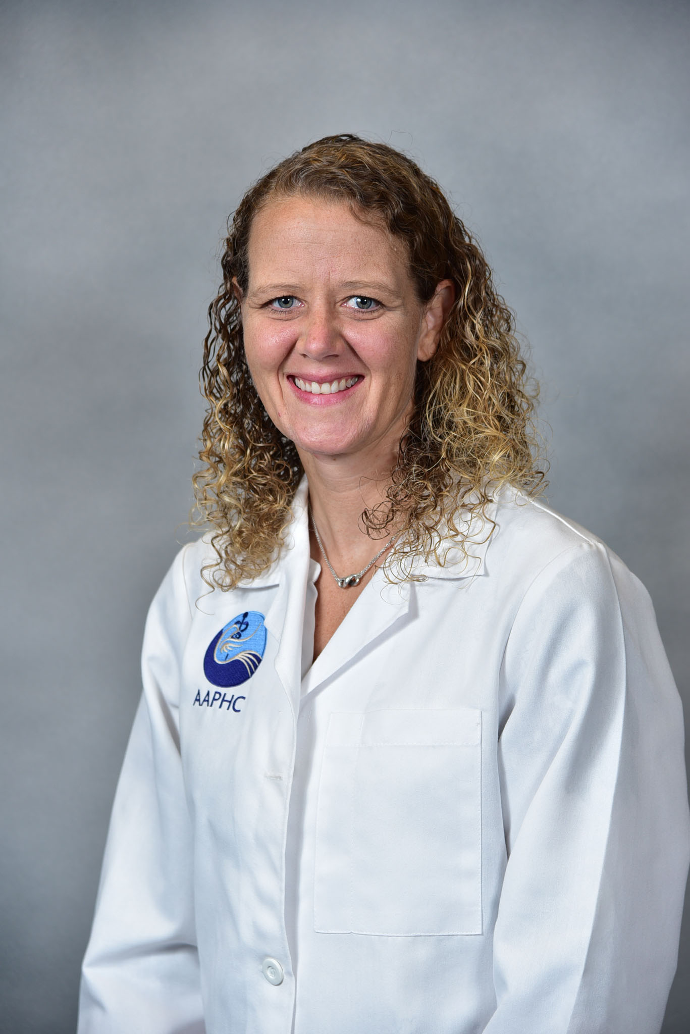 A woman with curly hair in a white lab coat smiles warmly in front of a gray background. A woman with curly hair in a white lab coat smiles warmly in front of a gray background.