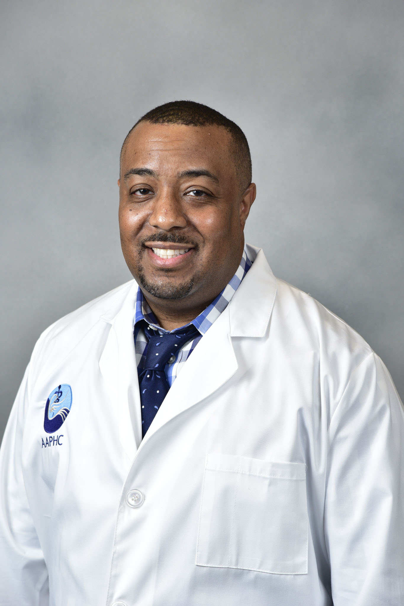 A smiling man in a white lab coat and blue tie stands against a plain gray background. A smiling man in a white lab coat and blue tie stands against a plain gray background.
