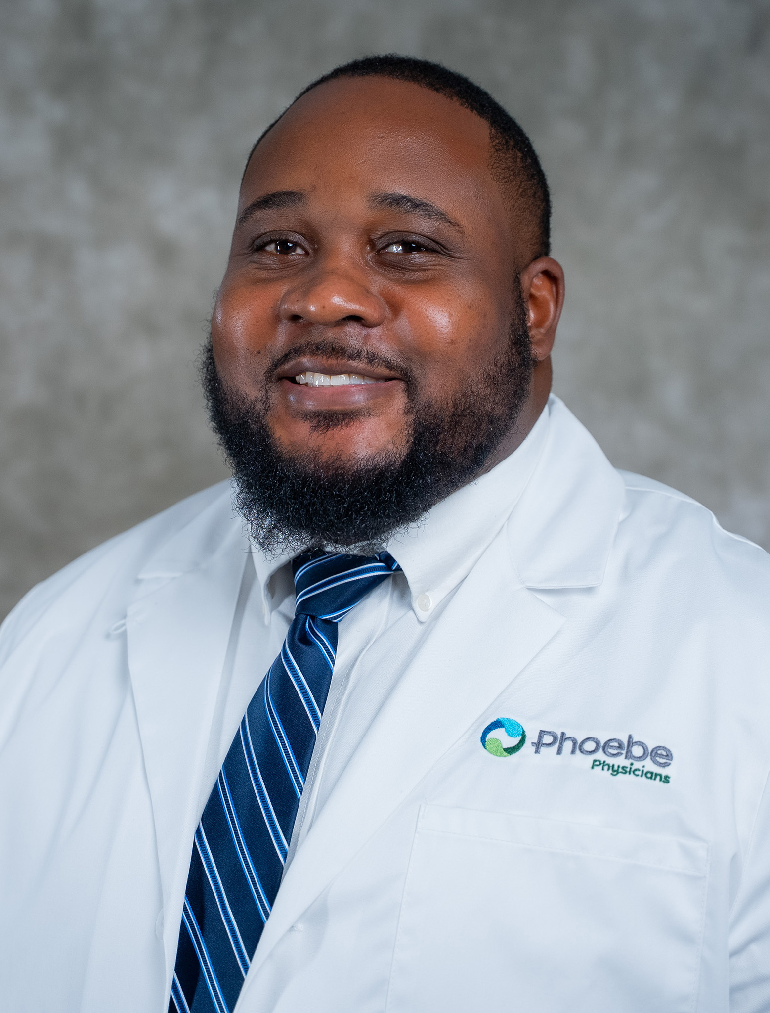 Smiling man with a beard in a white lab coat and striped tie stands before a gray background. Smiling man with a beard in a white lab coat and striped tie stands before a gray background.