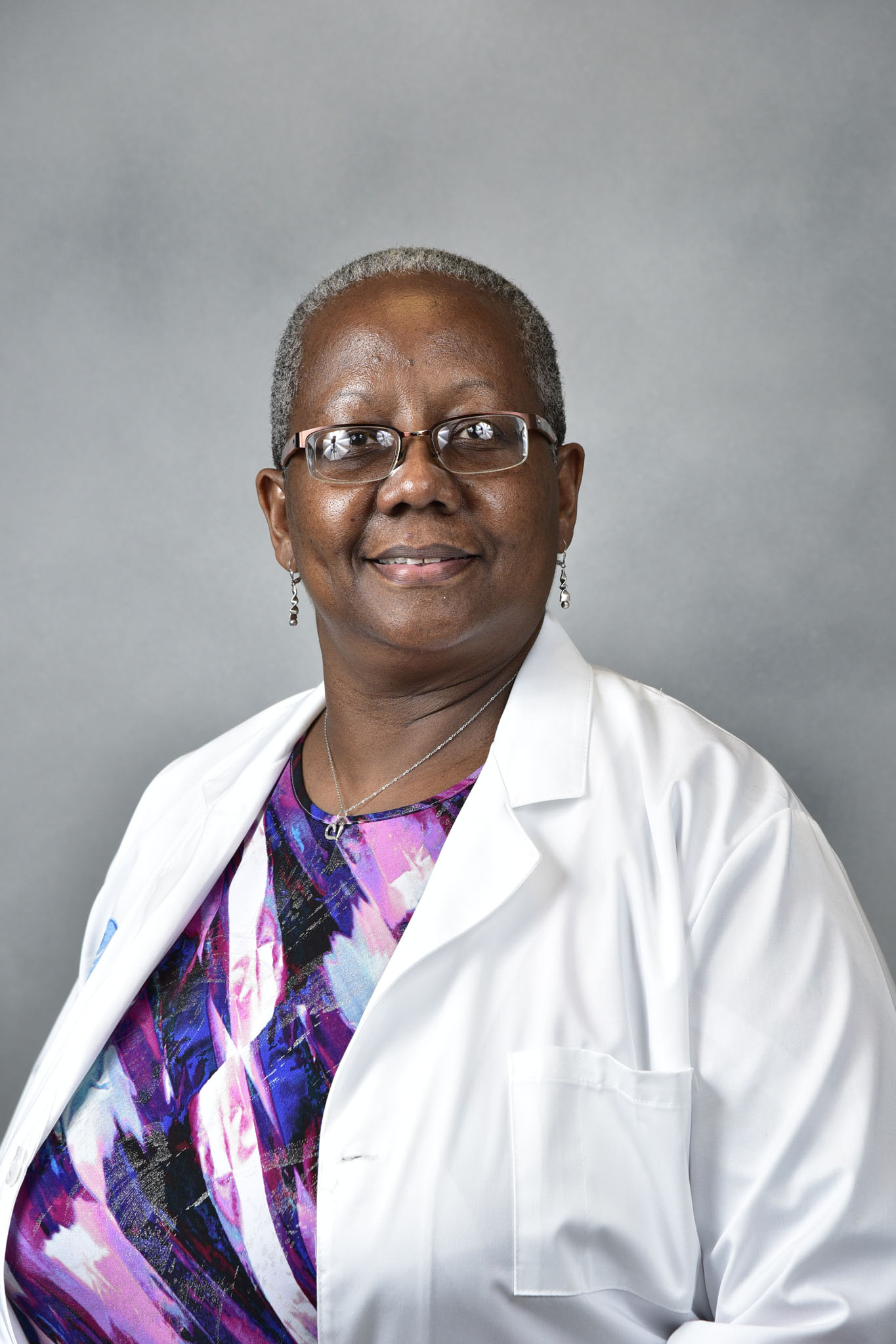 Smiling Deirdre Lawson with short hair, glasses, and a white lab coat over a colorful top stands against a gray background. Smiling Deirdre Lawson with short hair, glasses, and a white lab coat over a colorful top stands against a gray background.
