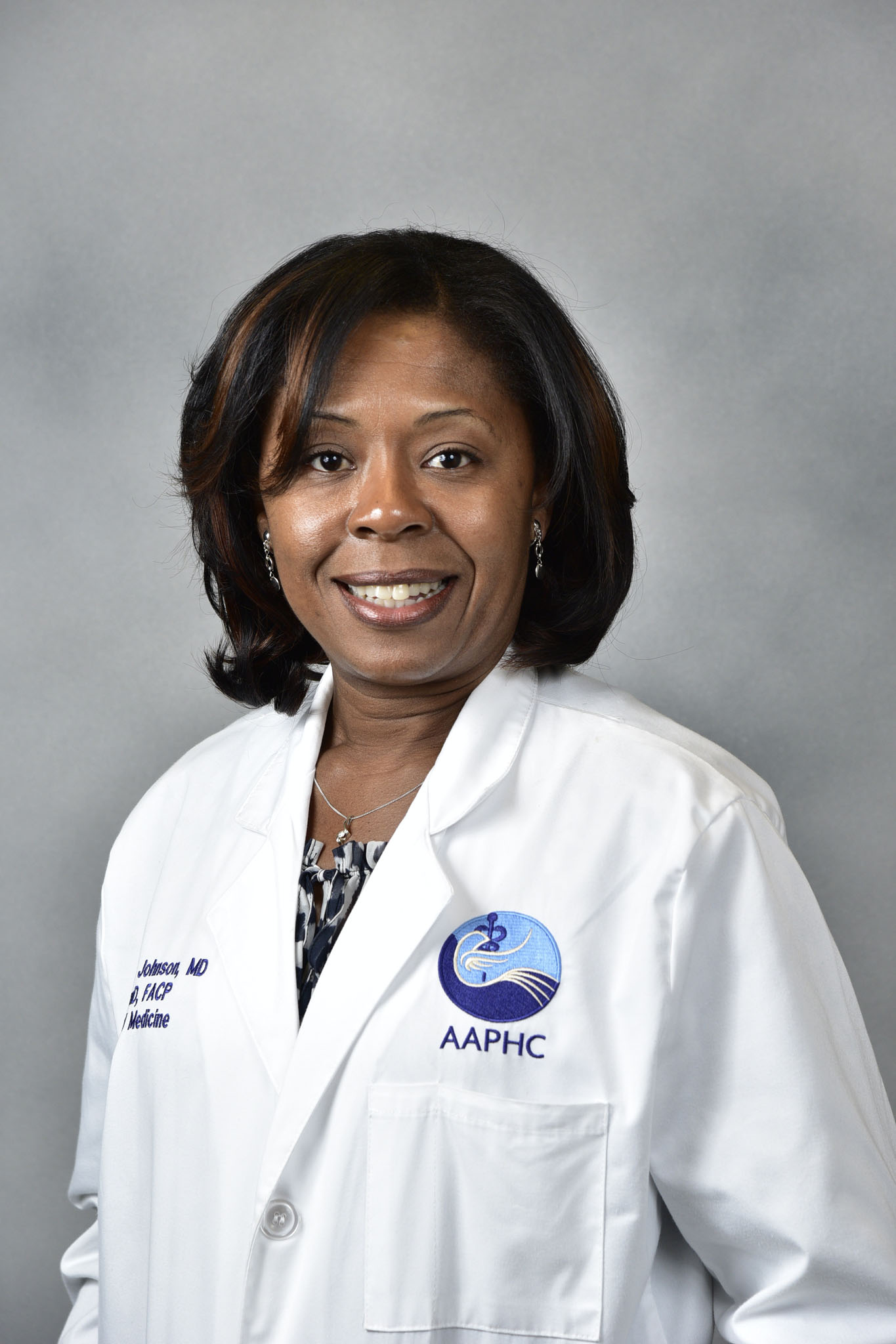 Smiling woman, Robyn Johnson, in a white medical coat with an AAPHC logo stands against a gray background. Smiling woman, Robyn Johnson, in a white medical coat with an AAPHC logo stands against a gray background.