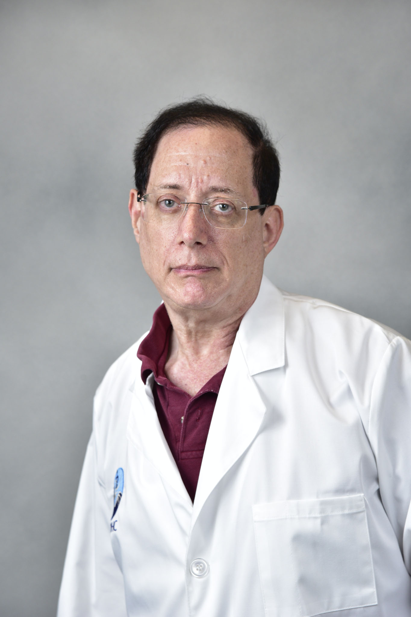 A person with short dark hair and glasses wears a white lab coat over a maroon shirt in front of a gray background. A person with short dark hair and glasses wears a white lab coat over a maroon shirt in front of a gray background.