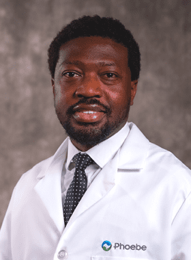 Babade_Oluwatomiwa A man with short curly hair and a beard, wearing a white lab coat with the logo Phoebe and a black dotted tie, poses against a neutral gray background.