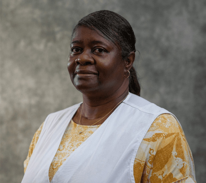 A woman with dark skin and black hair pulled back, wearing a yellow patterned top and a white vest, stands in front of a gray textured background, looking at the camera with a neutral expression.