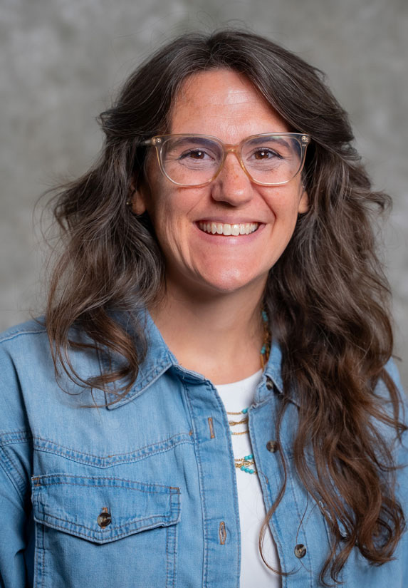 A smiling woman with long wavy brown hair and glasses wearing a light denim shirt over a white top, standing in front of a gray textured background.
