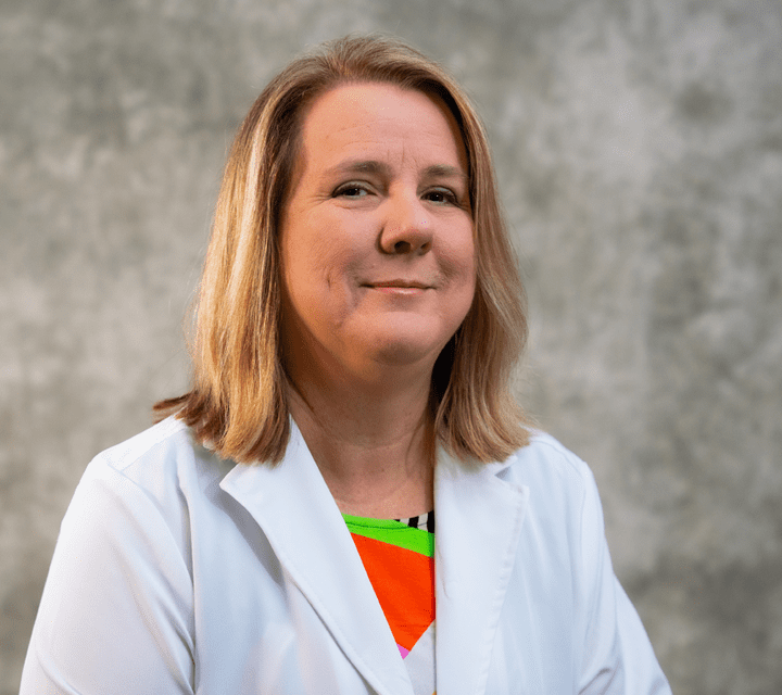 CherylEverson A woman with shoulder-length blonde hair wearing a white lab coat and colorful top, poses in front of a gray mottled background, smiling slightly at the camera.
