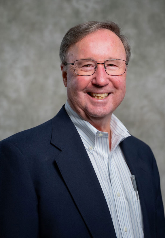 Smiling older man with glasses and gray hair wearing a dark blazer, striped shirt, and no tie, standing in front of a plain, light-colored background.