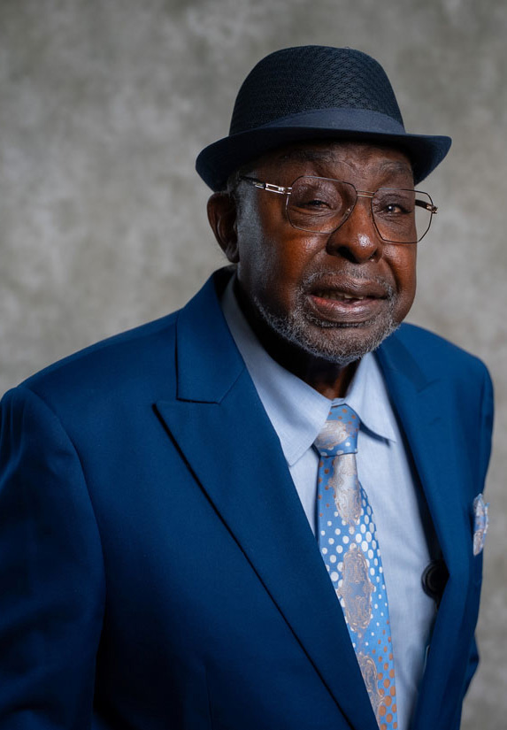 An elderly man wearing a blue suit, light blue shirt, patterned tie, and a dark blue hat poses in front of a mottled gray background, smiling slightly and wearing glasses.