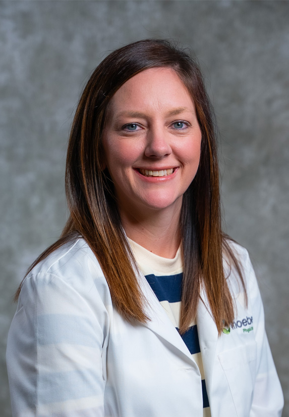 A woman with straight brown hair wearing a white lab coat and a striped shirt smiles at the camera in front of a gray background.