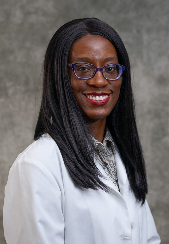 A woman wearing glasses and a white lab coat smiles in front of a gray background. She has long, straight black hair and is dressed professionally.