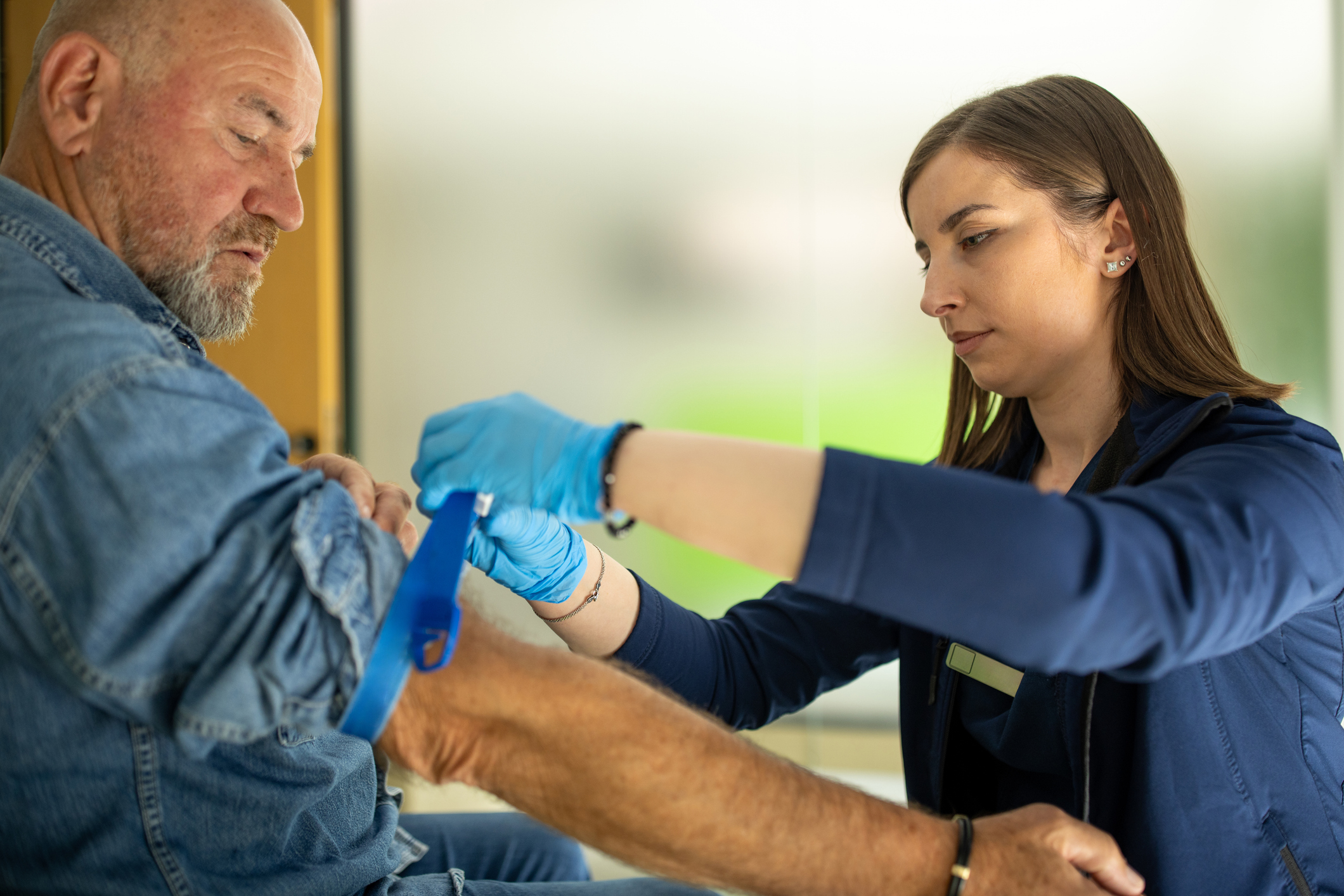 Nurse preparing a patient for a blood donation A healthcare worker wearing blue gloves applies a tourniquet to the arm of an older man in preparation for a medical procedure, possibly a blood draw or injection, in a clinical setting.