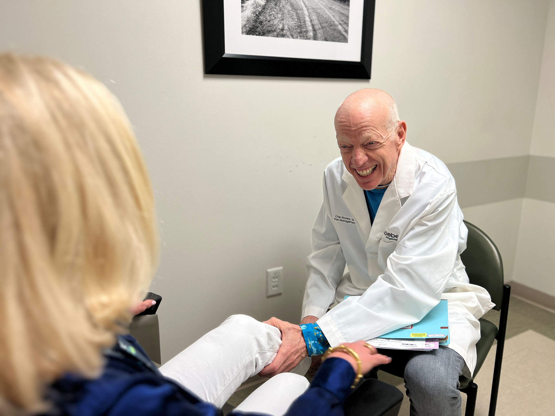 Pain Management A smiling doctor in a white coat examines a patient’s leg in a medical office, holding her ankle as they both engage in friendly conversation.