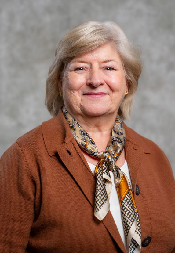 An older woman with short, light-colored hair is smiling, wearing a brown blazer and a patterned scarf, in front of a gray textured background.