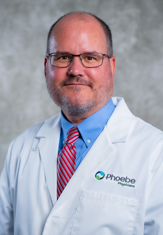 A man in glasses with a white lab coat, blue shirt, and red striped tie stands against a gray background. A man in glasses with a white lab coat, blue shirt, and red striped tie stands against a gray background.