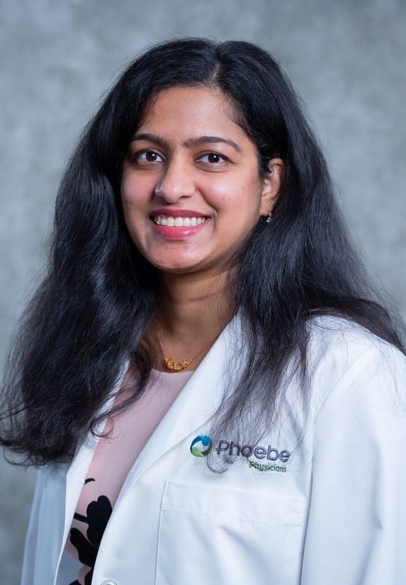 Dr. Shiny Teja Kolli, with long dark hair, smiles in a white coat with “Phoebe Physicians” logo against a gray background. Dr. Shiny Teja Kolli, with long dark hair, smiles in a white coat with "Phoebe Physicians" logo against a gray background.
