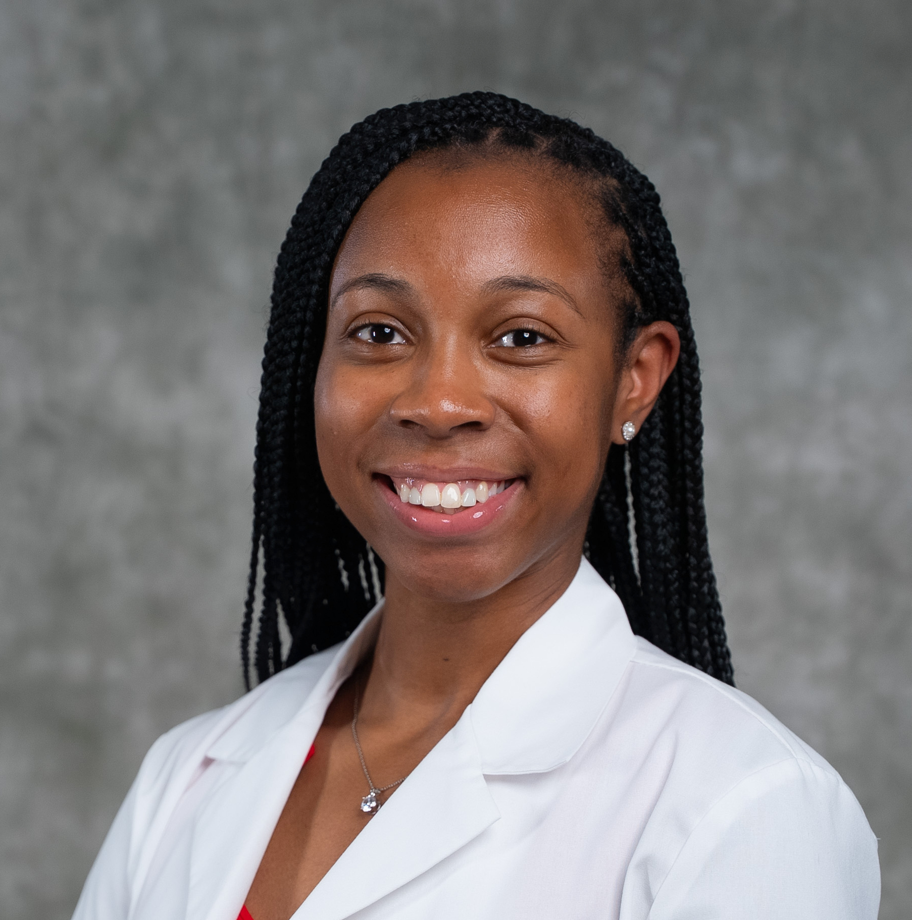 A woman with braided hair smiles, wearing a white lab coat and a necklace, standing against a gray background. A woman with braided hair smiles, wearing a white lab coat and a necklace, standing against a gray background.