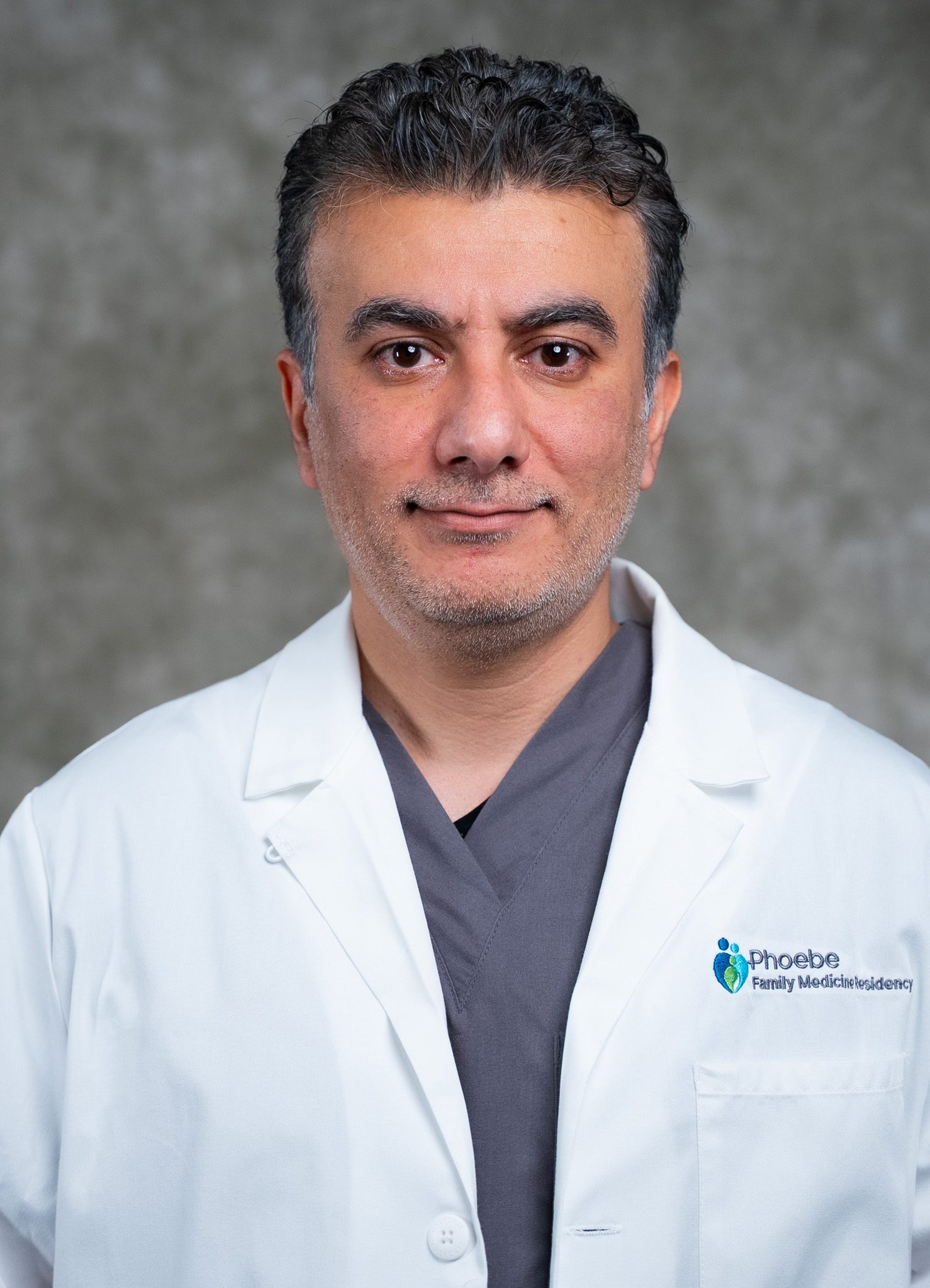 A man with short dark hair and a beard in a Phoebe Family Medicine Residency lab coat stands in front of a light gray background. A man with short dark hair and a beard in a Phoebe Family Medicine Residency lab coat stands in front of a light gray background.