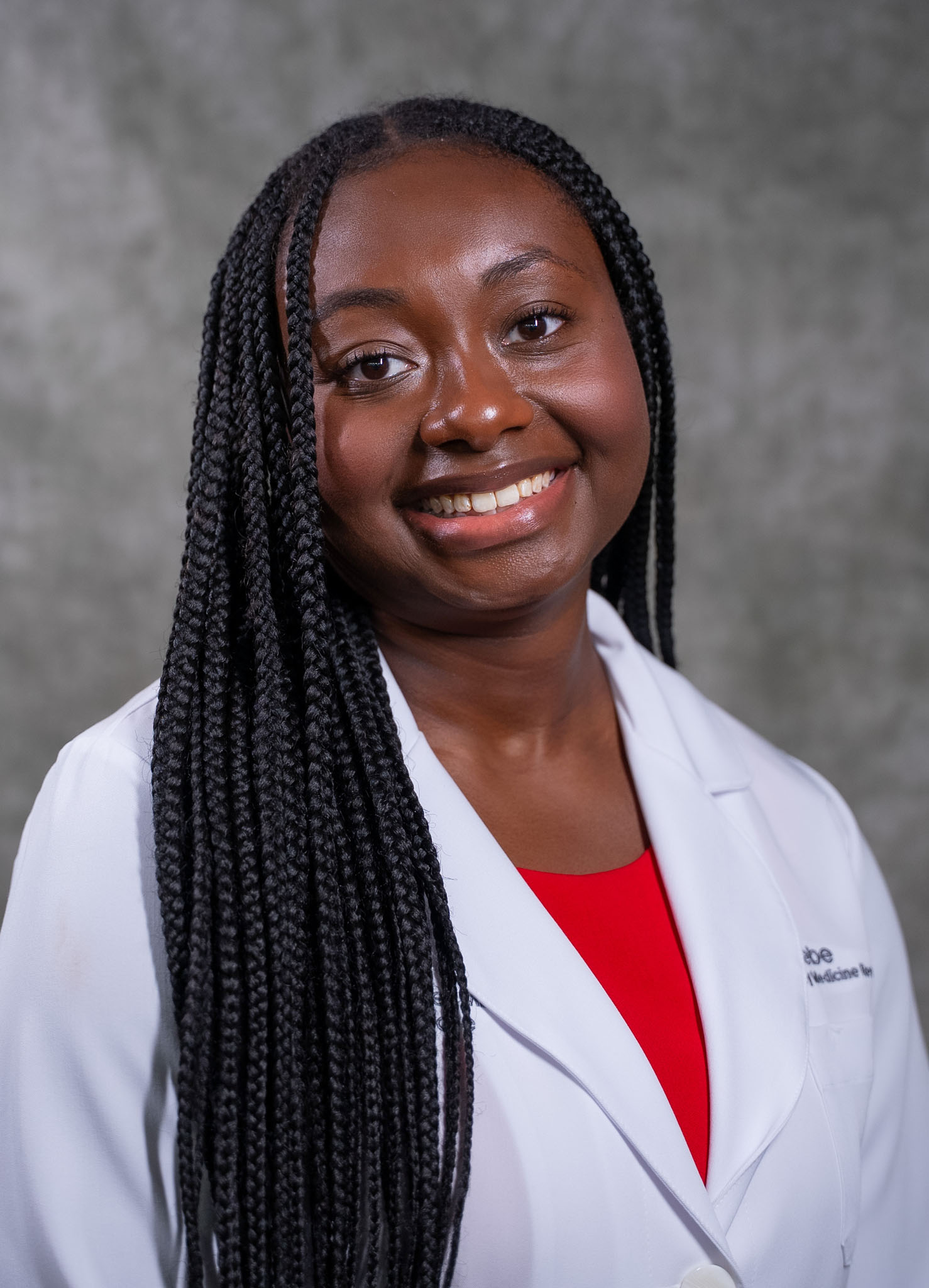 A woman in a white lab coat and red top smiles in front of a gray background. A woman in a white lab coat and red top smiles in front of a gray background.