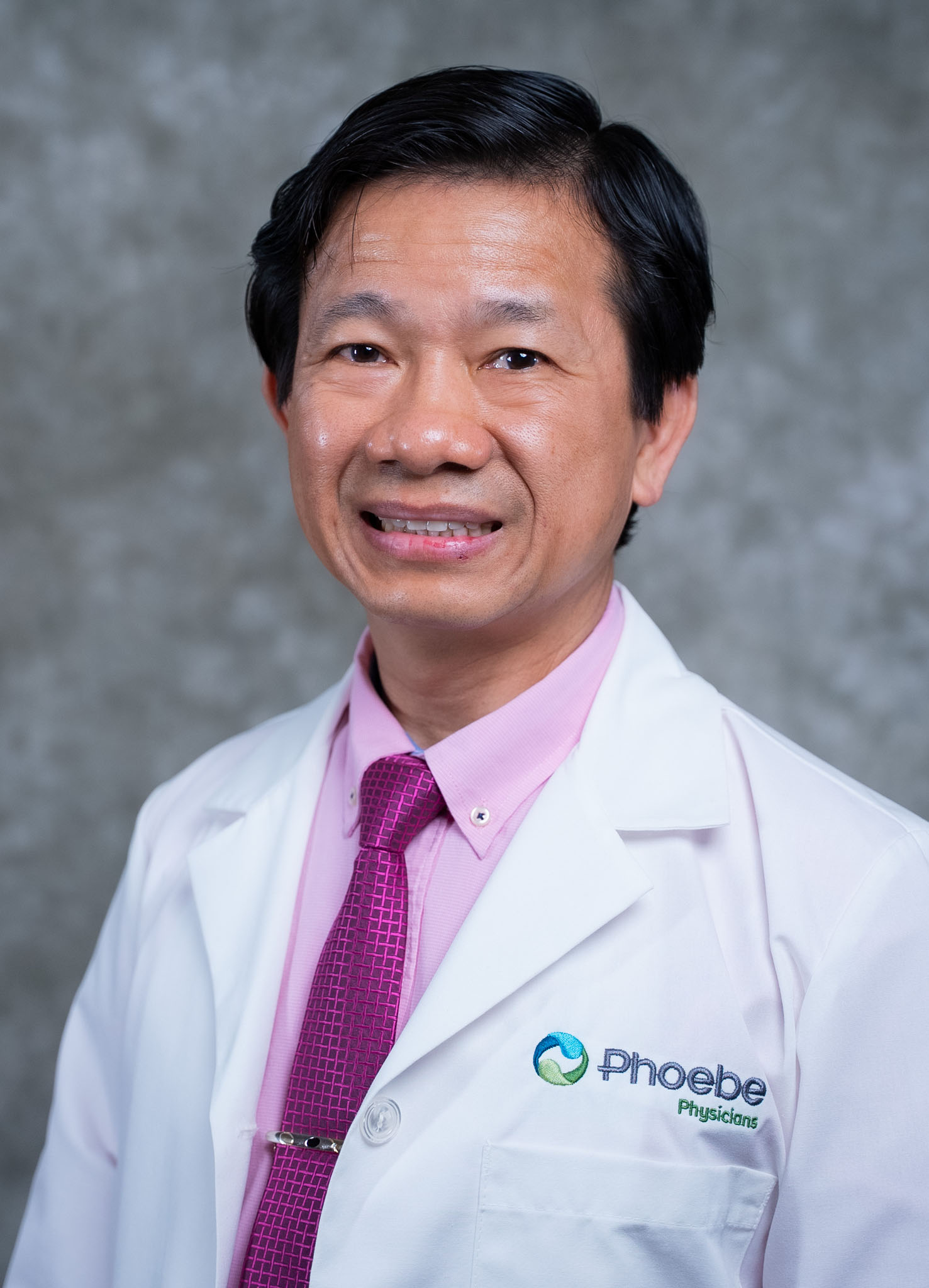 Man in a white lab coat and pink shirt, smiling, with Physicians group logo on his coat, gray background.