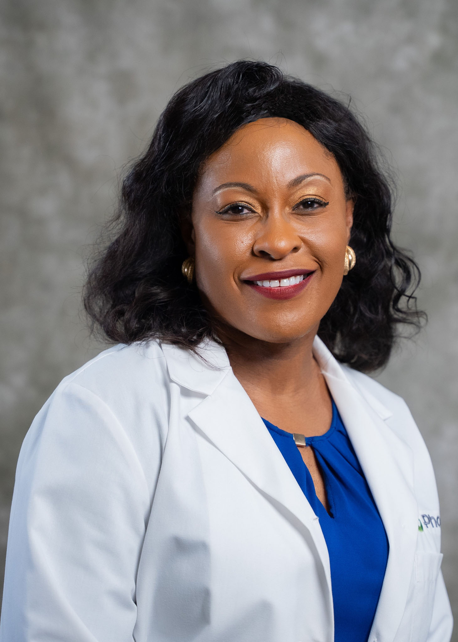 A person in a white lab coat and blue blouse smiles at the camera against a gray background.