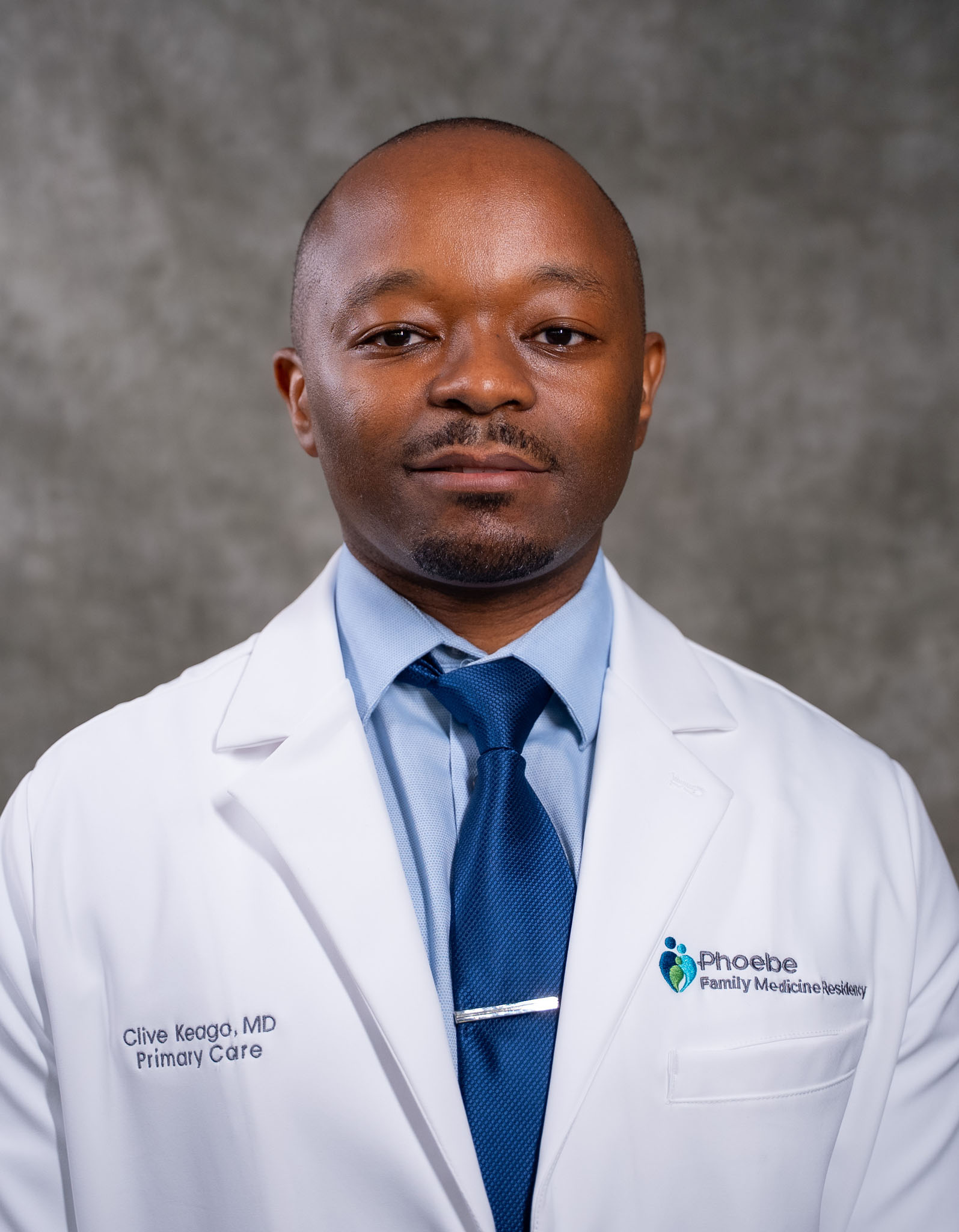 A man in a white coat and blue tie stands against a plain gray background, looking directly at the camera. A man in a white coat and blue tie stands against a plain gray background, looking directly at the camera.