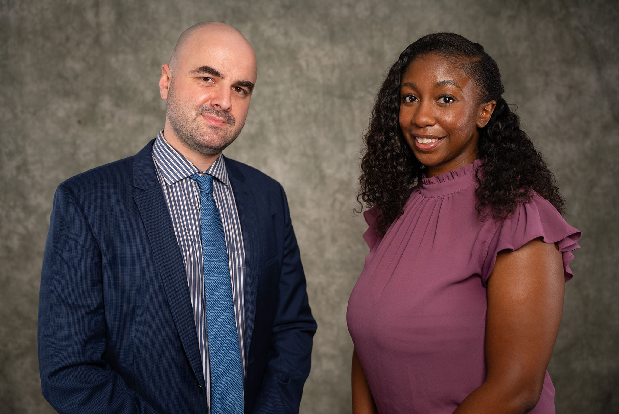 A man in a suit and a woman in a purple top smile together in front of a gray, textured backdrop. A man in a suit and a woman in a purple top smile together in front of a gray, textured backdrop.
