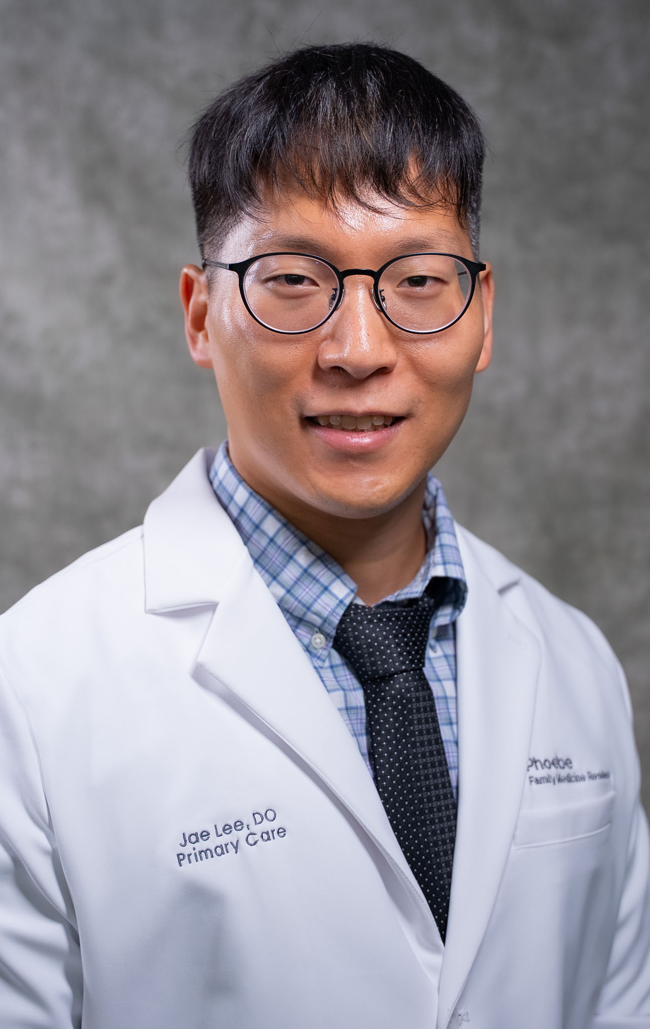 A man wearing glasses and a white doctor’s coat smiles in front of a gray background. A man wearing glasses and a white doctor’s coat smiles in front of a gray background.