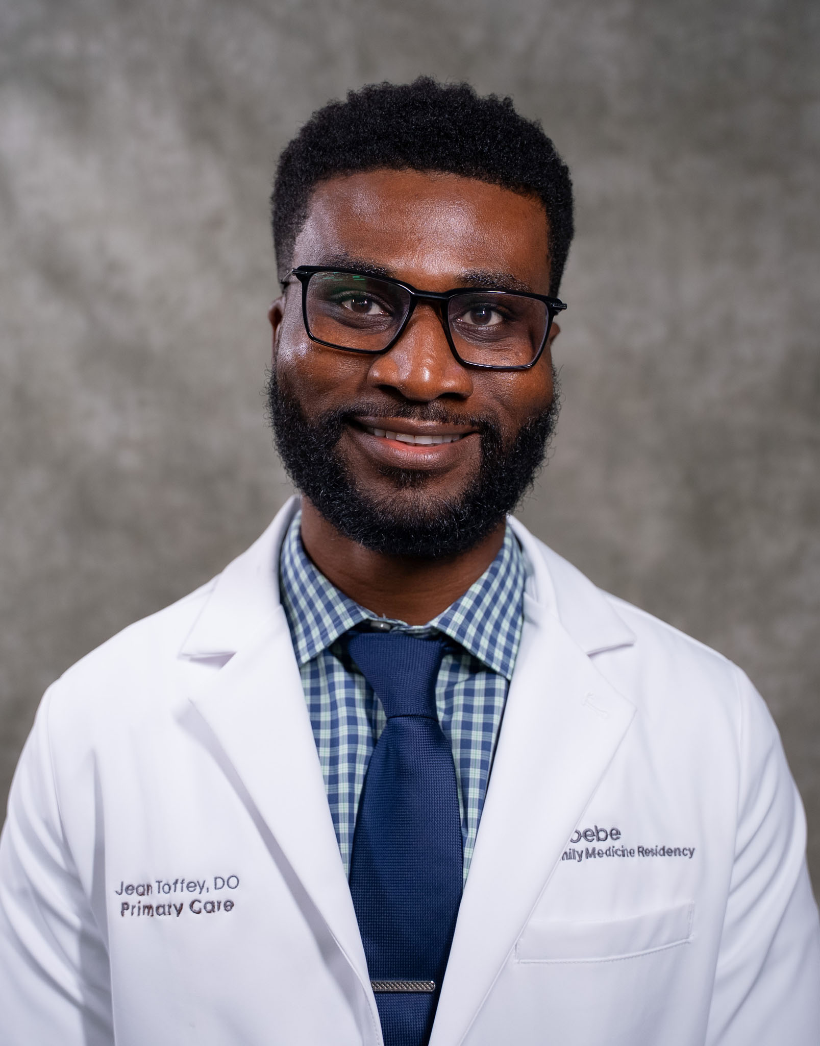 A man in a white lab coat, glasses, and a blue tie smiles at the camera against a gray background. A man in a white lab coat, glasses, and a blue tie smiles at the camera against a gray background.