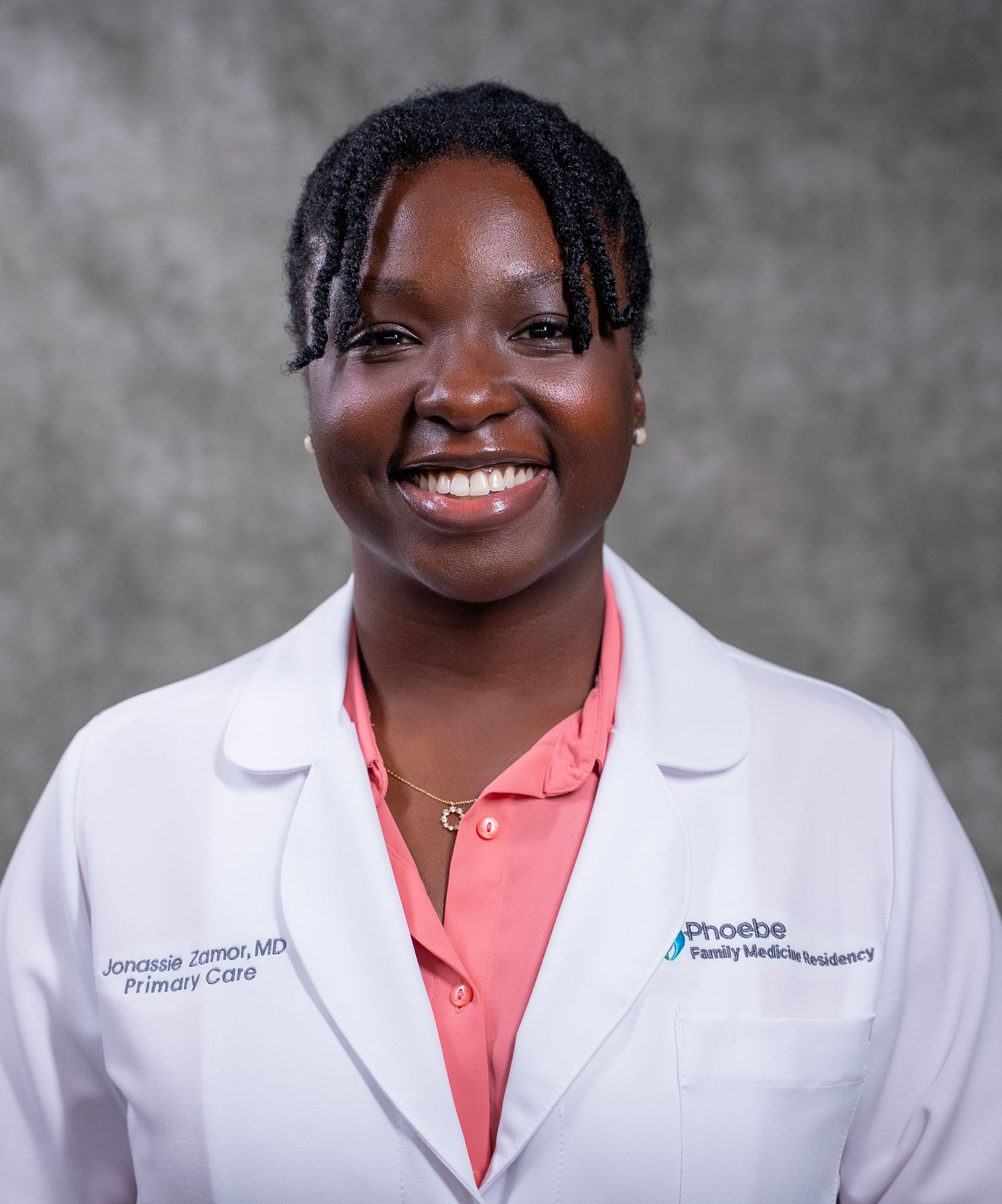 A woman in a white lab coat and pink shirt smiles while standing in front of a gray background. A woman in a white lab coat and pink shirt smiles while standing in front of a gray background.