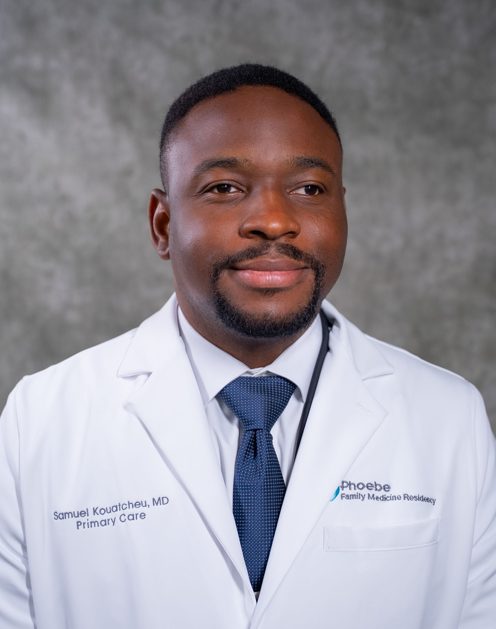 A man wearing a white doctor’s coat and tie smiles while standing in front of a plain gray background. A man wearing a white doctor’s coat and tie smiles while standing in front of a plain gray background.
