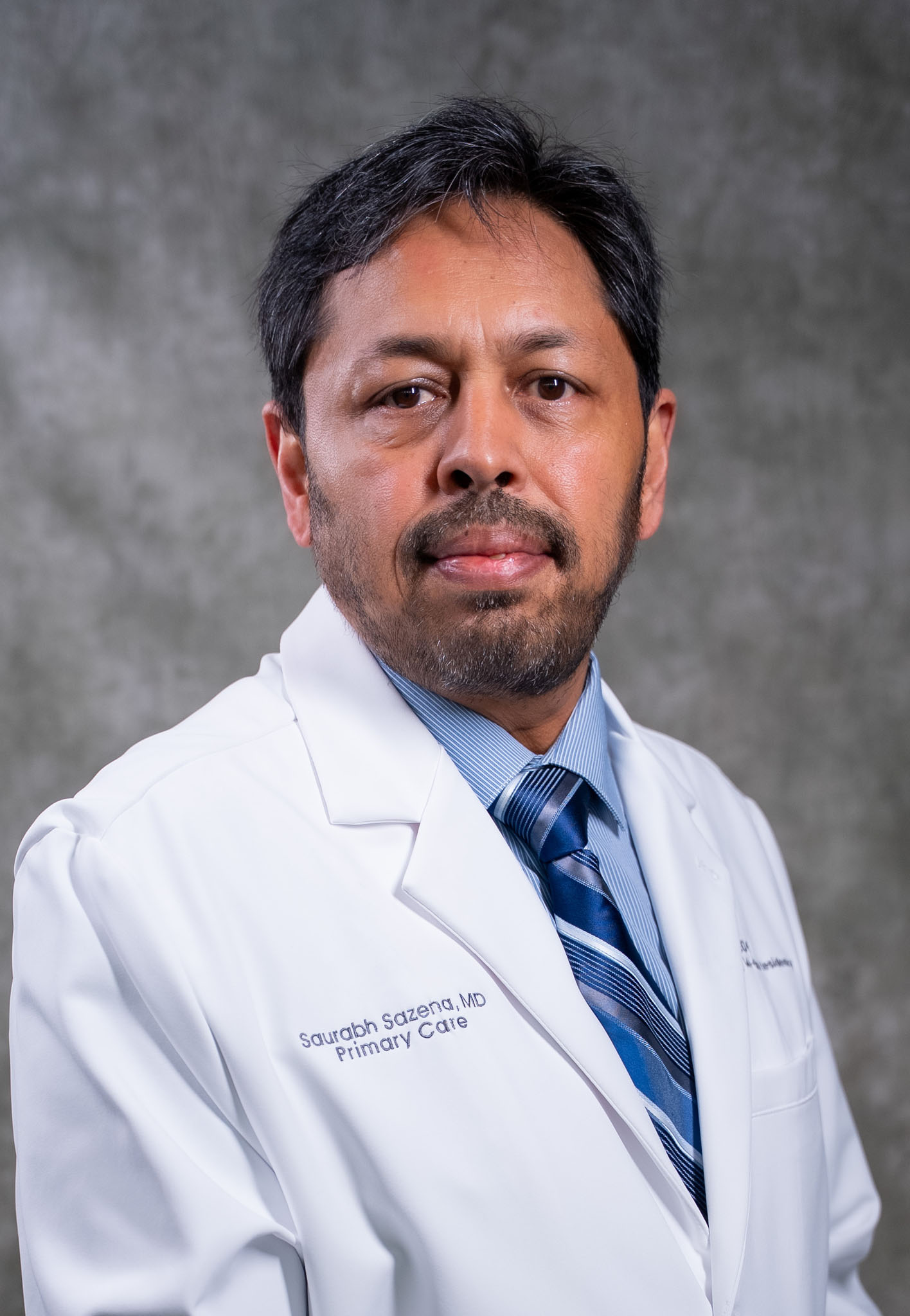 Man with short dark hair and beard wearing a white lab coat and blue striped tie, posing against a gray background. Man with short dark hair and beard wearing a white lab coat and blue striped tie, posing against a gray background.