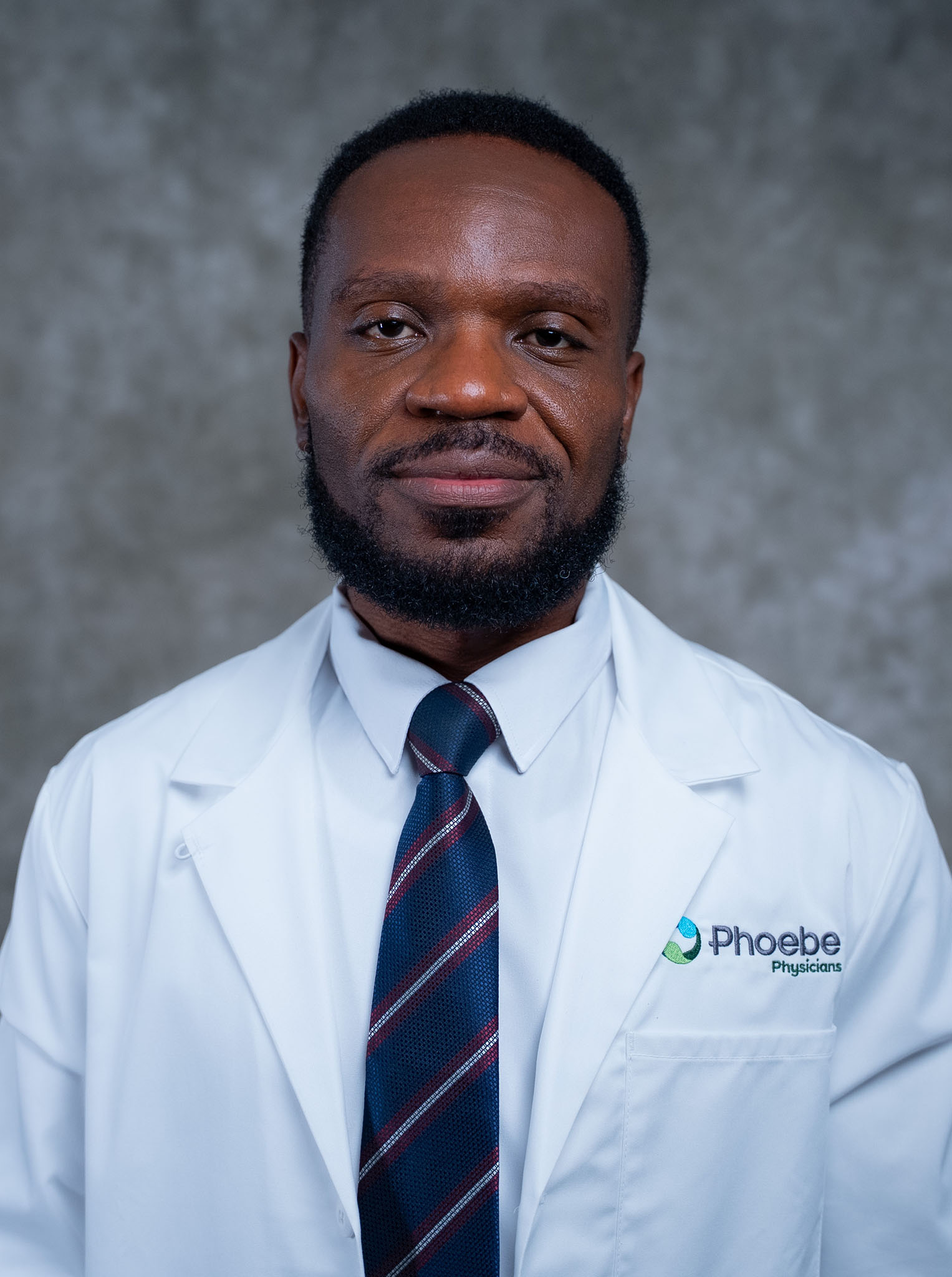 A person in a white lab coat and tie stands before a gray background, wearing a slight smile. A person in a white lab coat and tie stands before a gray background, wearing a slight smile.
