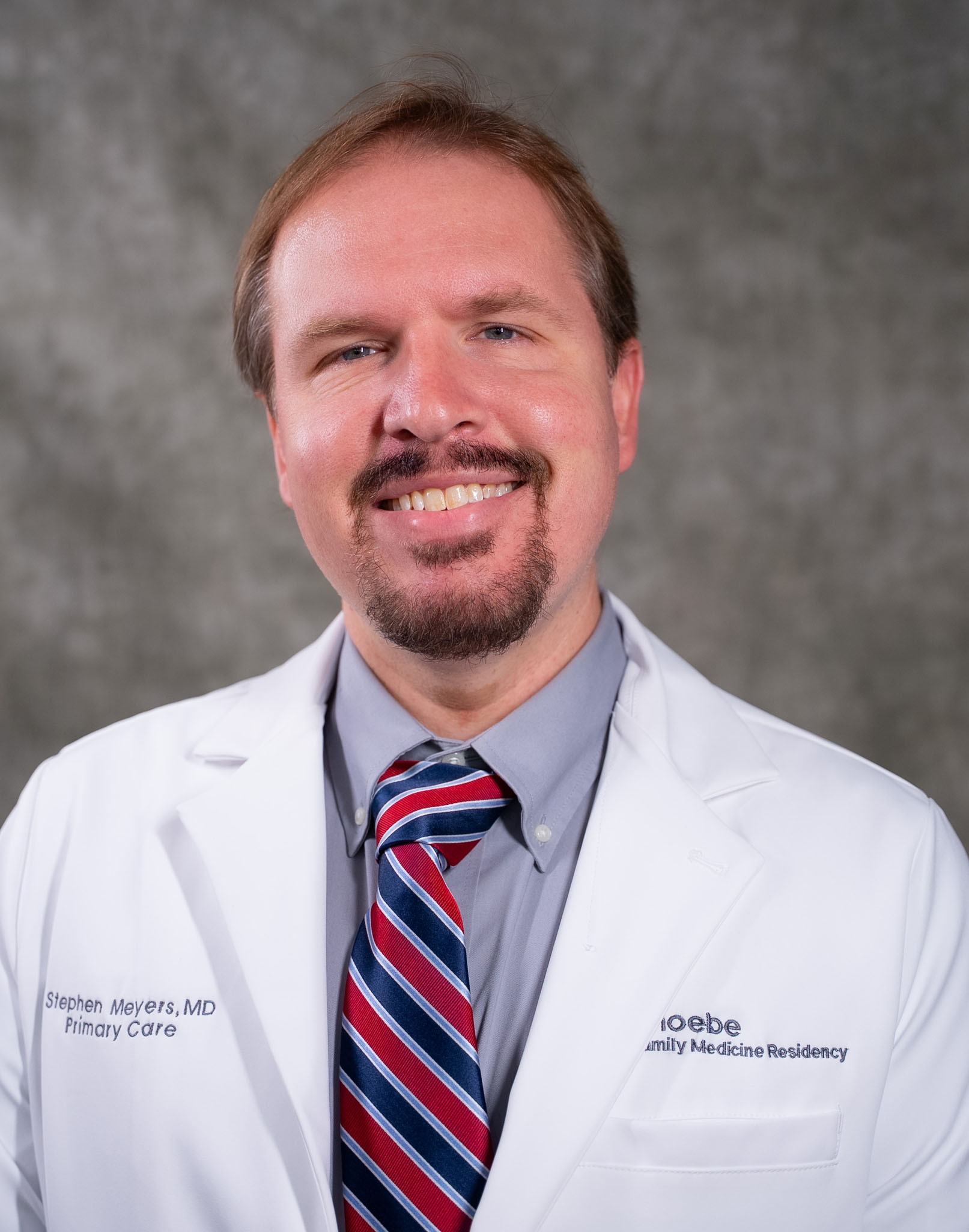 A smiling man in a white lab coat and striped tie stands against a gray background. A smiling man in a white lab coat and striped tie stands against a gray background.