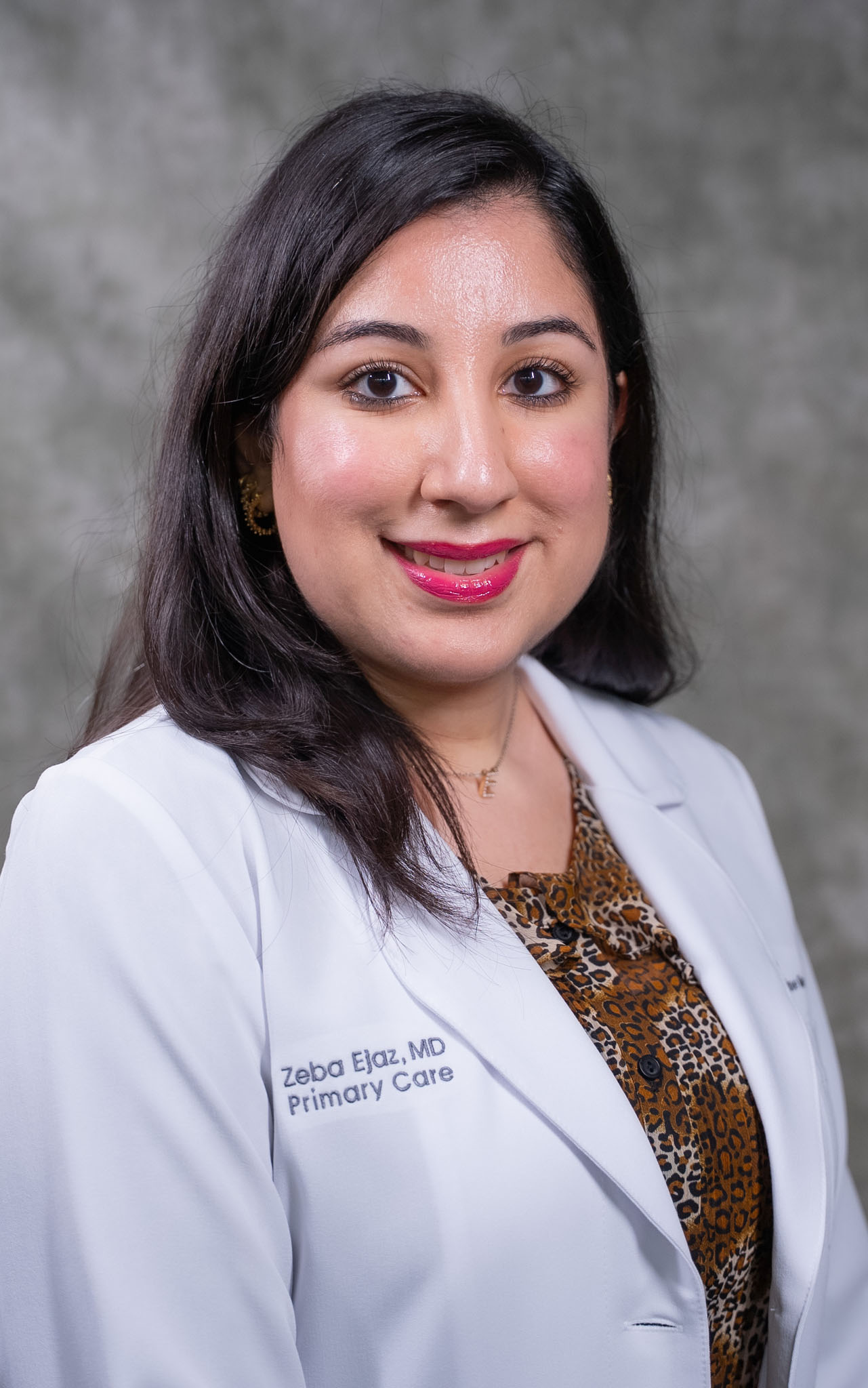 A woman in a white lab coat smiles in front of a gray background. Her coat reads Zeba Ejaz, MD, Primary Care. A woman in a white lab coat smiles in front of a gray background. Her coat reads Zeba Ejaz, MD, Primary Care.