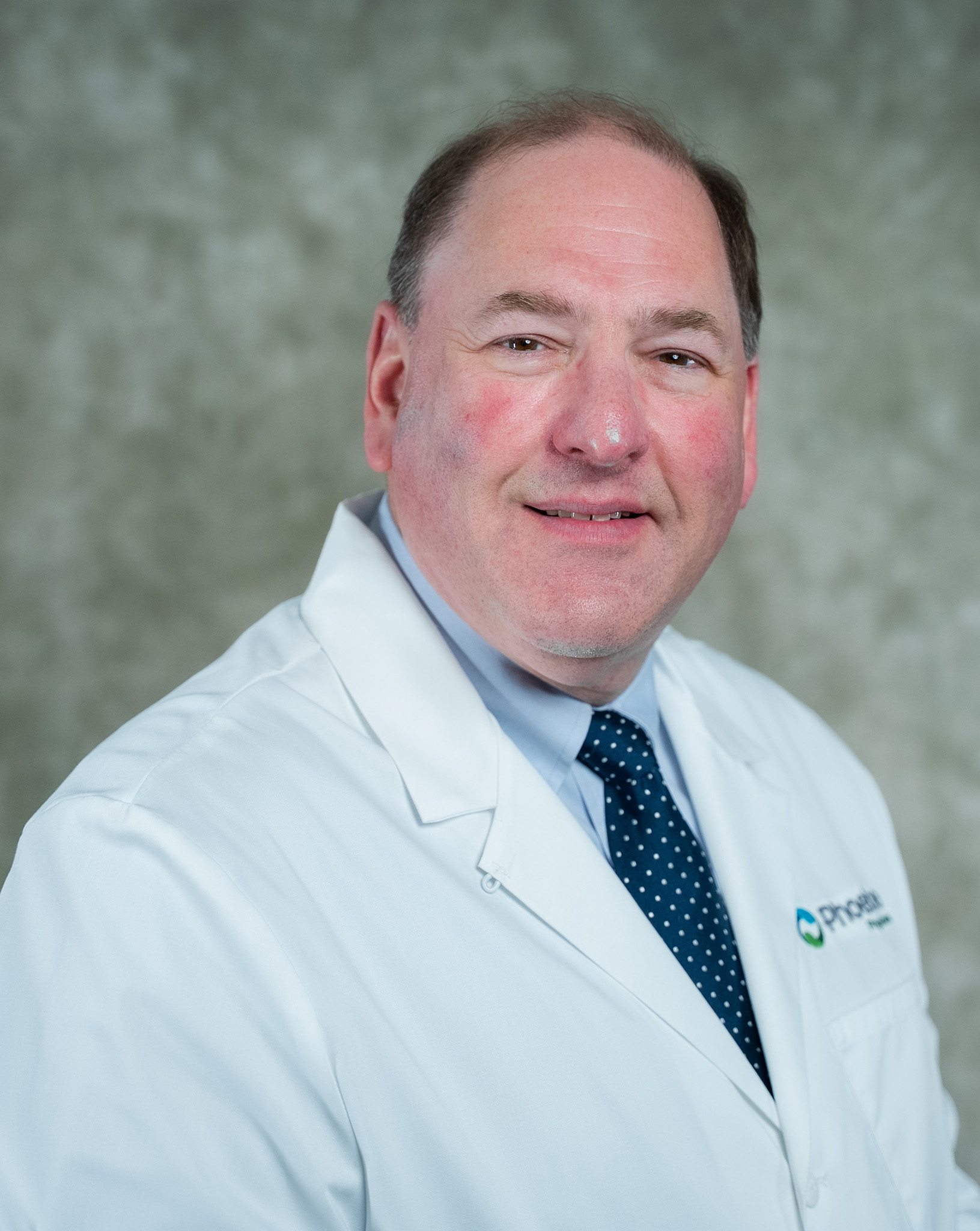 A man in a white lab coat and navy blue polka dot tie poses against a gray backdrop. A man in a white lab coat and navy blue polka dot tie poses against a gray backdrop.