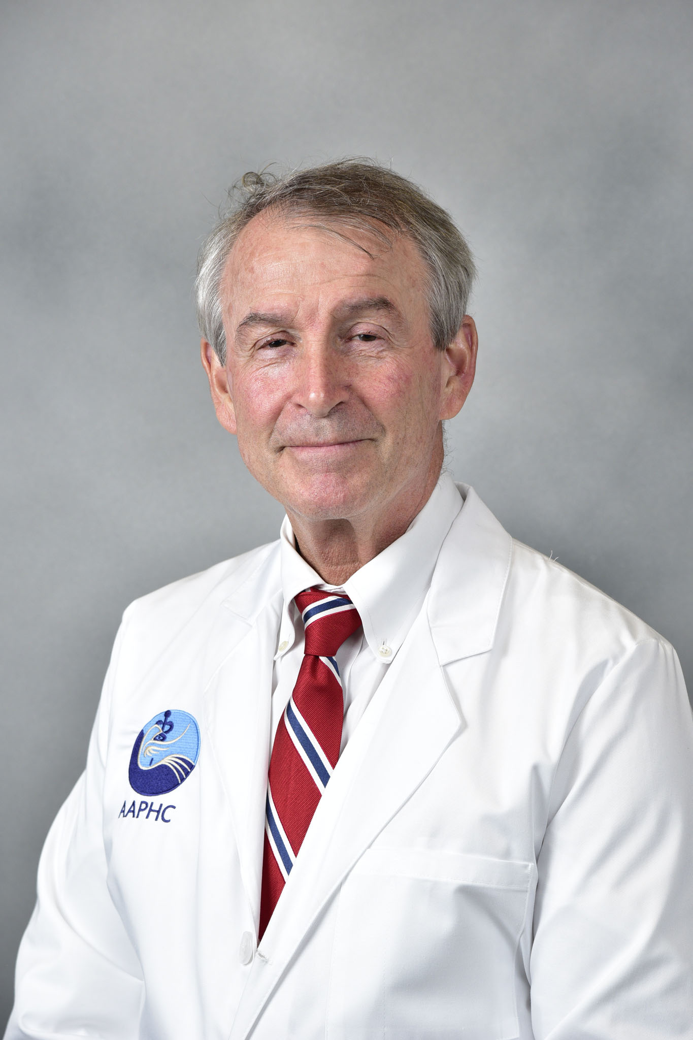 Hotz A James A man with short brown hair wearing a white doctors coat, a blue shirt, and a red tie stands in front of a green background, smiling slightly at the camera.
