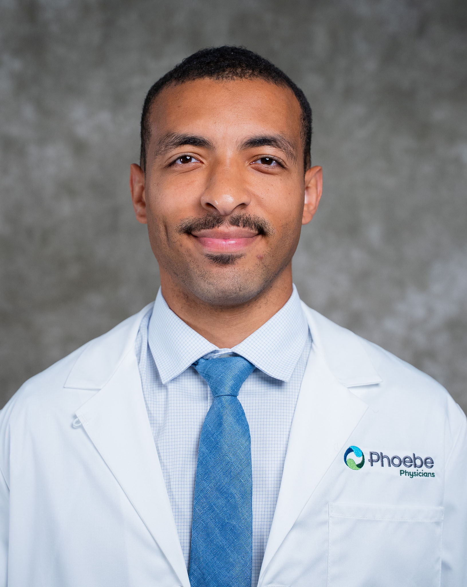 Man in a white lab coat and blue tie smiling against a gray background. Christopher Creighton, Phoebe Physicians logo visible. Man in a white lab coat and blue tie smiling against a gray background. Christopher Creighton, Phoebe Physicians logo visible.