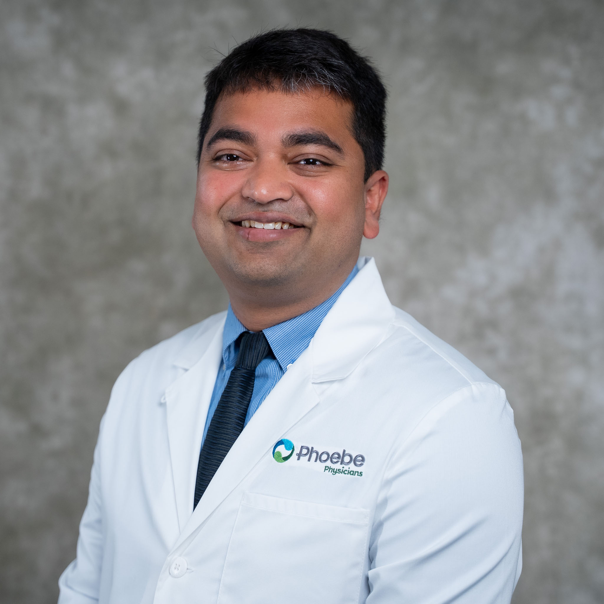 Smiling man in a white lab coat and tie, standing in front of a gray mottled background. Smiling man in a white lab coat and tie, standing in front of a gray mottled background.