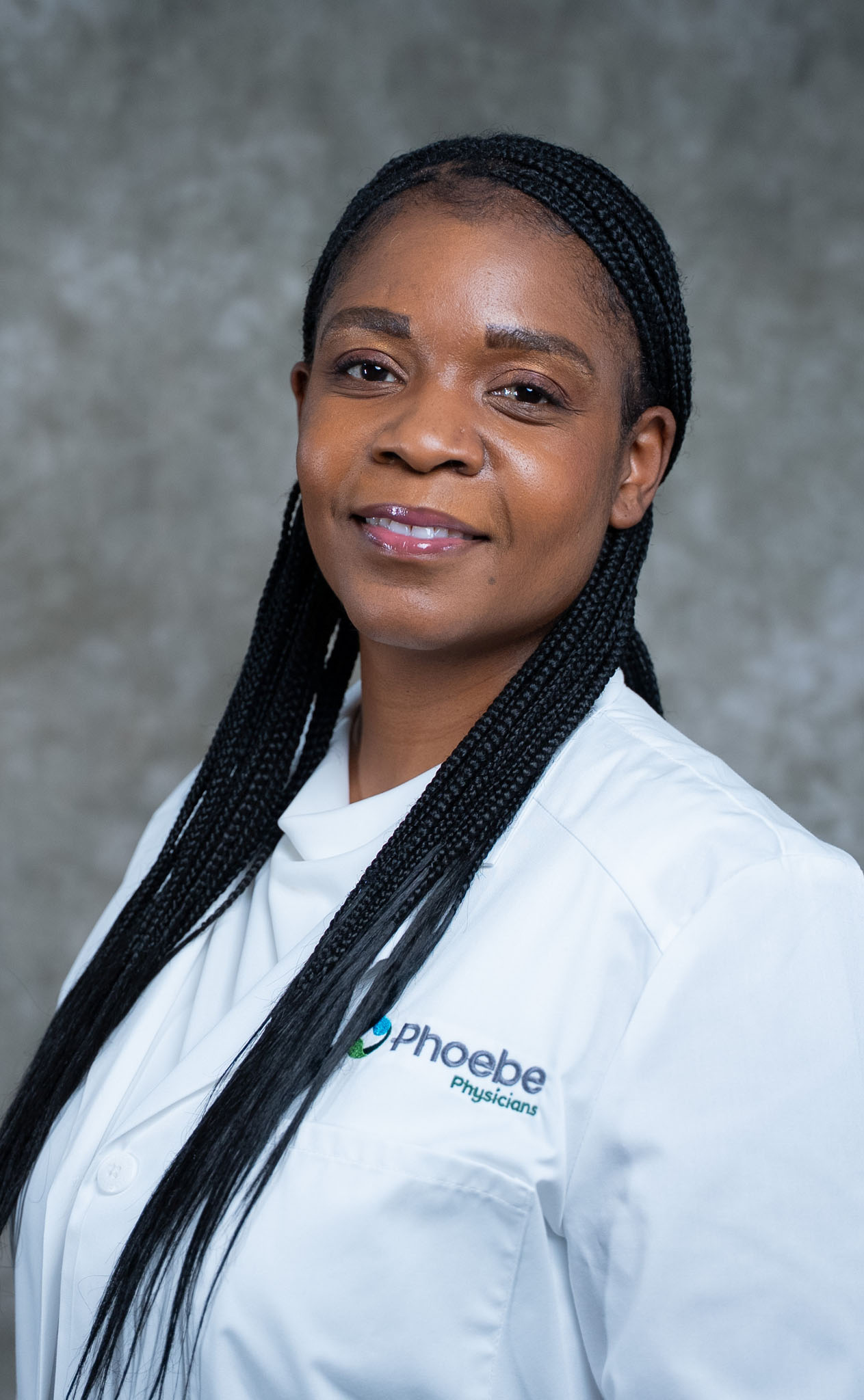 Kethia Phelizor, wearing a white medical coat with the Phoebe Physicians logo, poses confidently against a gray background. Kethia Phelizor, wearing a white medical coat with the Phoebe Physicians logo, poses confidently against a gray background.