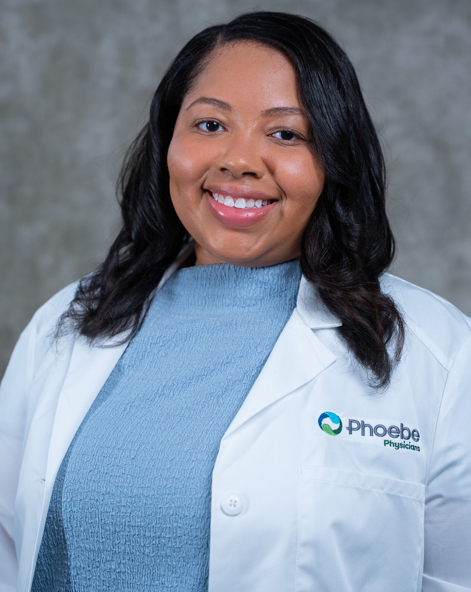 Sylvette Rogers, in a white doctor’s coat featuring the Phoebe Physicians logo, smiles warmly against a gray background. Sylvette Rogers, in a white doctor’s coat featuring the Phoebe Physicians logo, smiles warmly against a gray background.