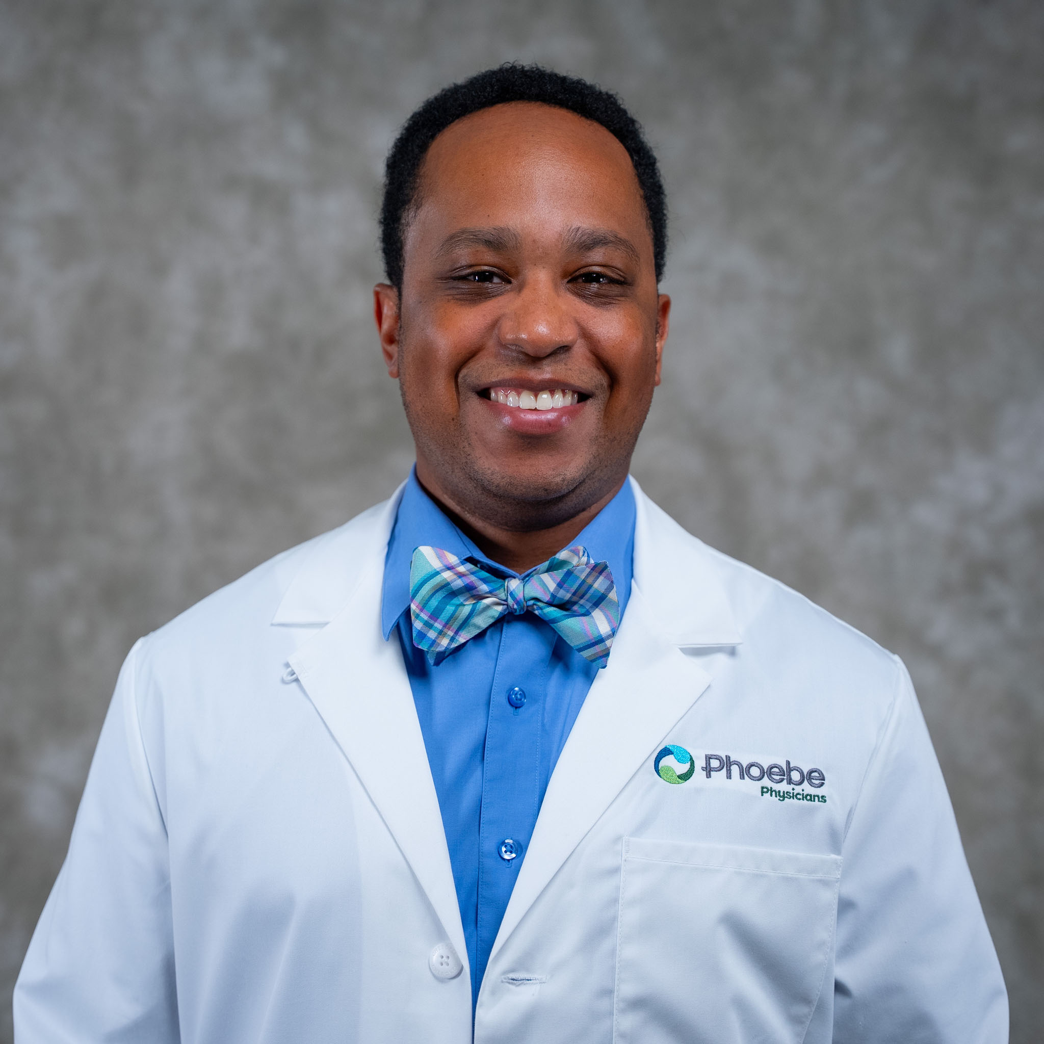 A smiling man in a white lab coat and bow tie stands against a gray background. A smiling man in a white lab coat and bow tie stands against a gray background.