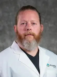 A man with a reddish-brown beard and mustache, wearing a white medical coat over a dark shirt, poses in front of a gray textured background.