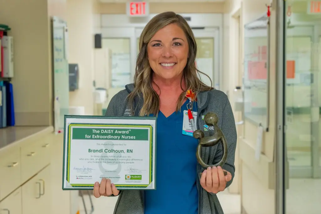 A smiling Phoebe Nurse stands in a hospital hallway holding a DAISY Award certificate and trophy, celebrating her achievement in Compassionate NICU Care.