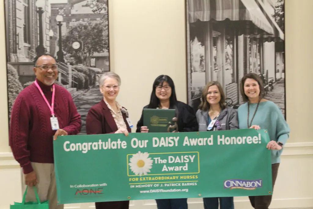 Five people stand indoors, smiling and holding a green banner that reads Congratulate Our DAISY Award Honoree! at Phoebe Sumter Medical Center. They celebrate compassionate nursing care as Tomomi Colquitt holds a certificate in the center.