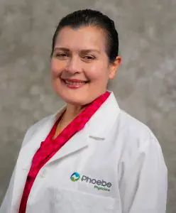 Maria Jasinskas, a woman with dark hair pulled back, wearing a white lab coat embroidered with Phoebe Physicians and a bright pink patterned blouse, smiles in front of a gray textured background.