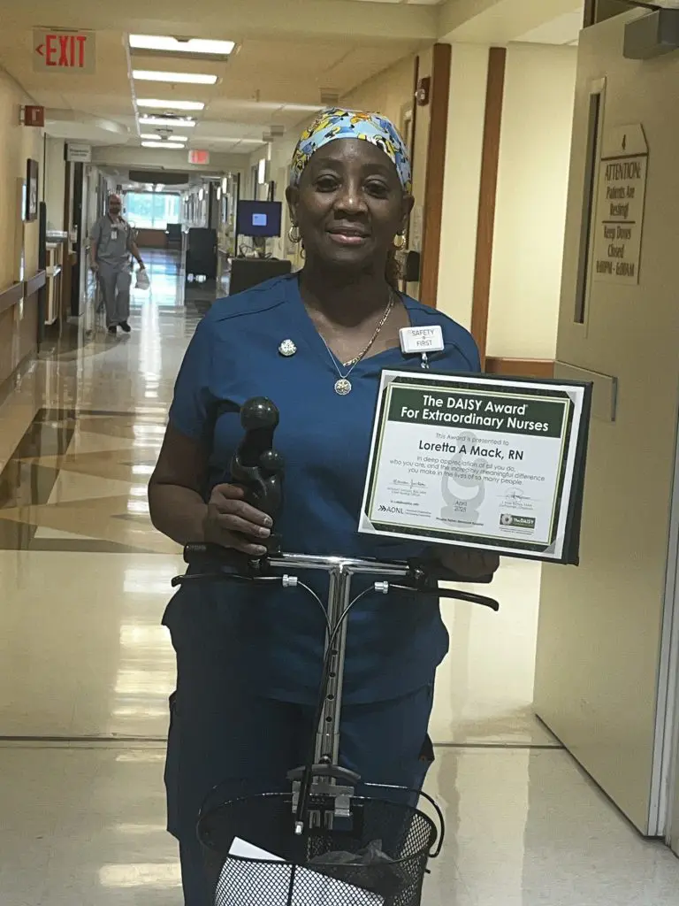 A Phoebe Nurse in blue scrubs and a colorful headscarf stands in a hallway, proudly holding a DAISY Award certificate and trophy, smiling at the camera in celebration of nursing recognition.