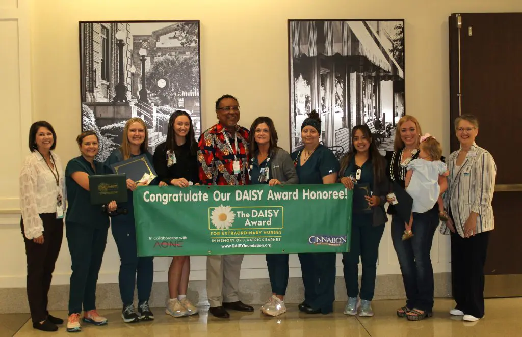 A group of people, including several ICU nurses in scrubs, stand indoors holding a green banner that reads Congratulations Our DAISY Award Honoree! in celebration of exceptional care. Some are smiling, and one person holds a certificate.