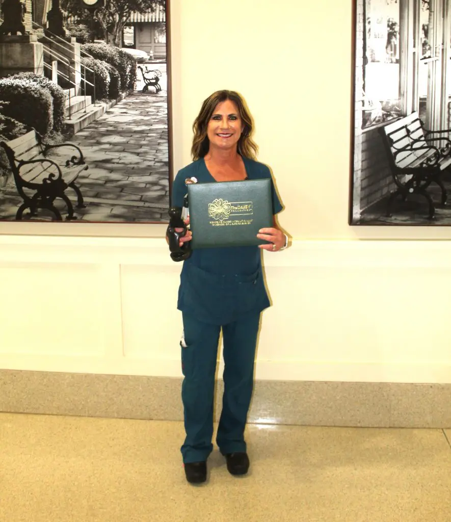 A Pre-Op Nurse in teal medical scrubs stands indoors, smiling and holding a cordless drill. Black and white photos of benches and buildings hang on the wall behind her at PSMC.