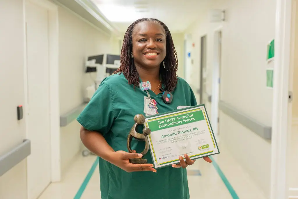 A Labor & Delivery Nurse in green scrubs smiles while holding a DAISY Award certificate and a trophy in a PPMH hospital hallway.