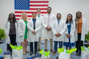 Eight people in white lab coats pose and smile indoors, celebrating Phoebe’s new family medicine residents, gift bags in front.